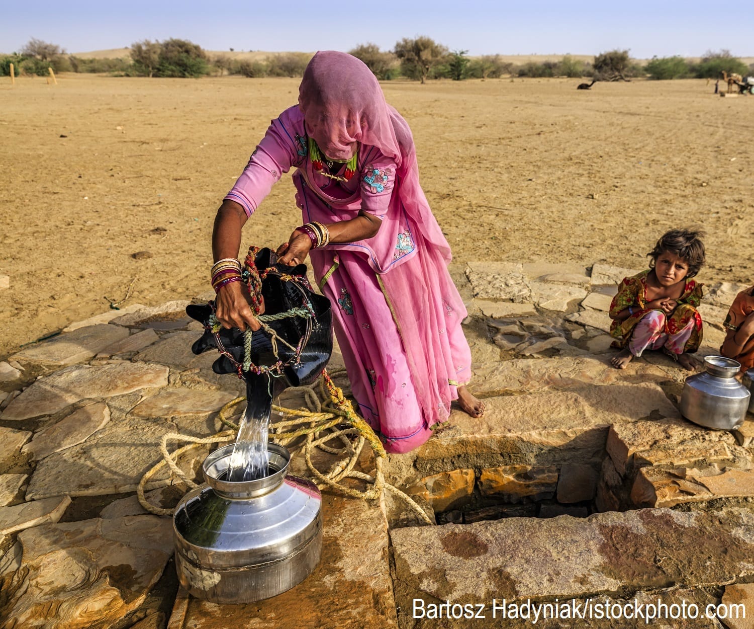 An Indian woman drawing water from a well while her children wait beside her.