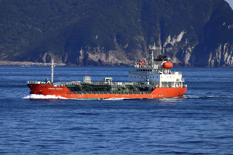 Orange and white chemical tanker moves through ocean against mountain backdrop