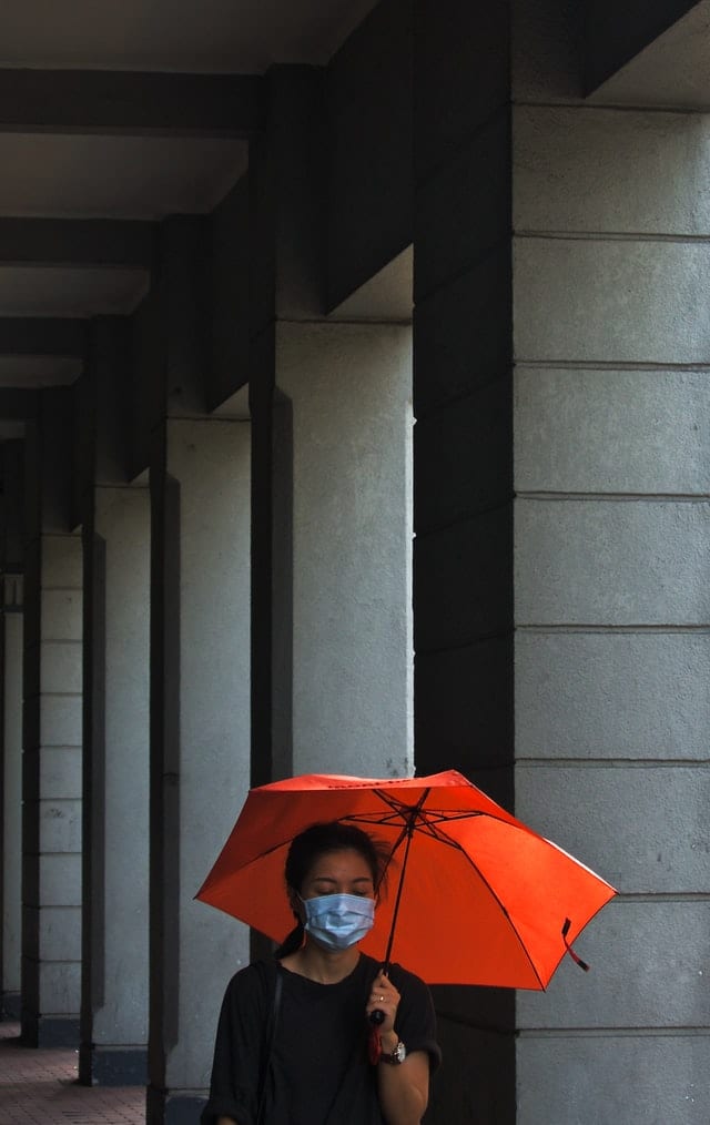 Woman in black shirt walks by columns while wearing a medical mask and holding an orange umbrella