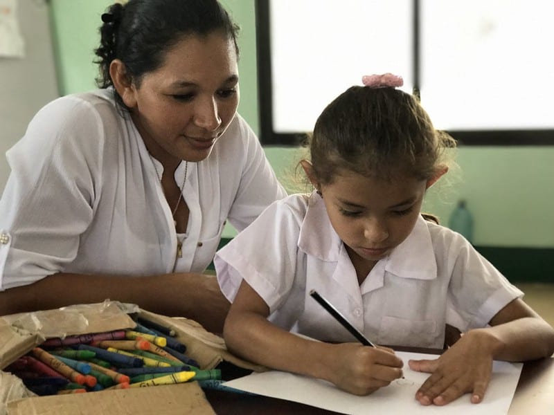 Female preschool student writes at desk while female teacher looks over her shoulder