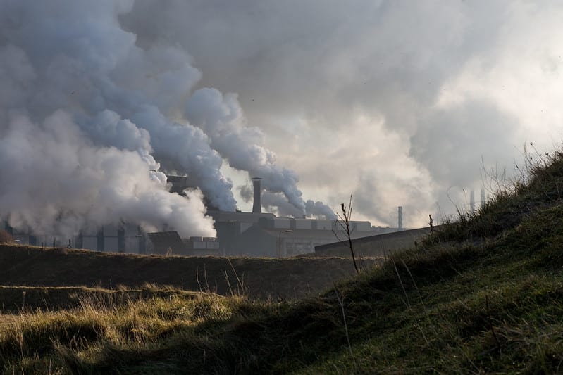 Factories behind grassy hills billow clouds of smoke that cover the sky