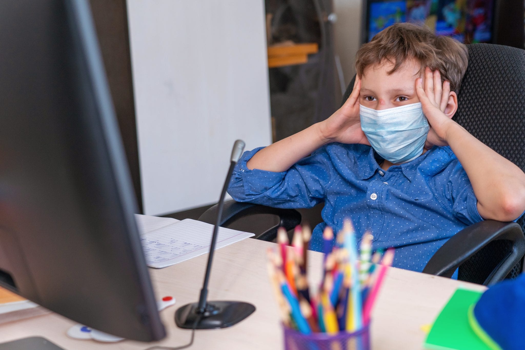 Student wearing a mask and sitting at a desk, holds hands to his head in frustration while staring at a computer screen