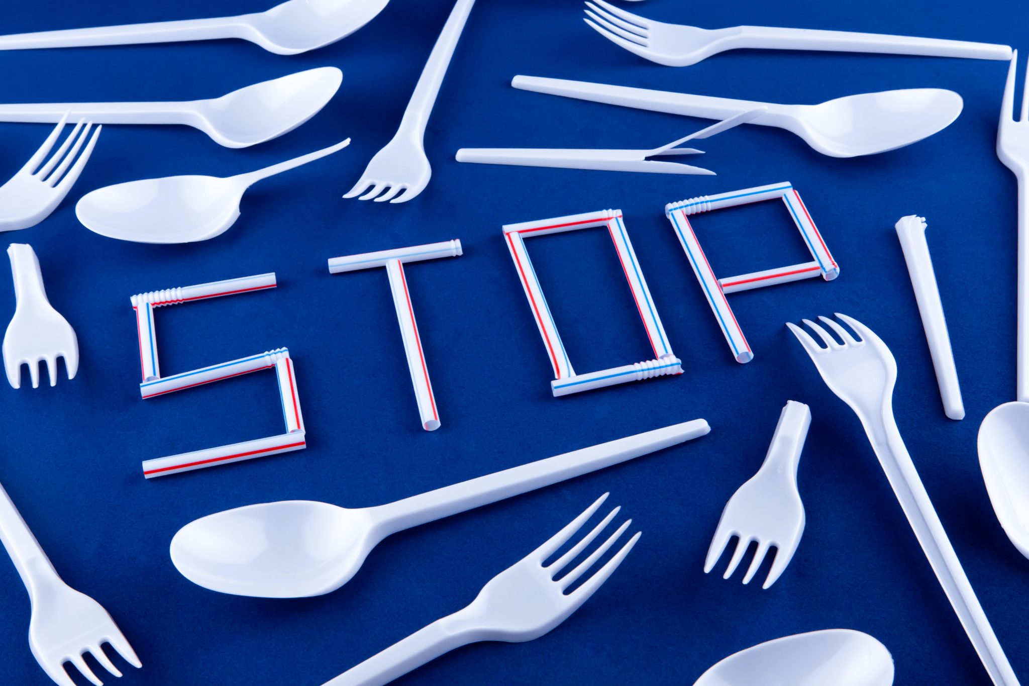 Plastic forks and spoons are scattered atop a blue surface, surrounding plastic straws that spell out the word "stop"
