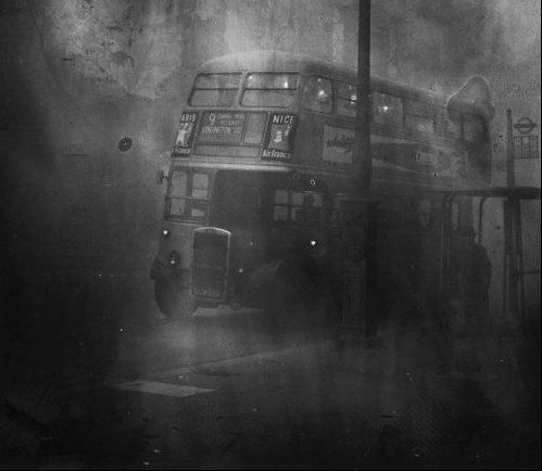 A 1950s black and white photo shows a double decker bus parked on a London street in a cloud of smog.