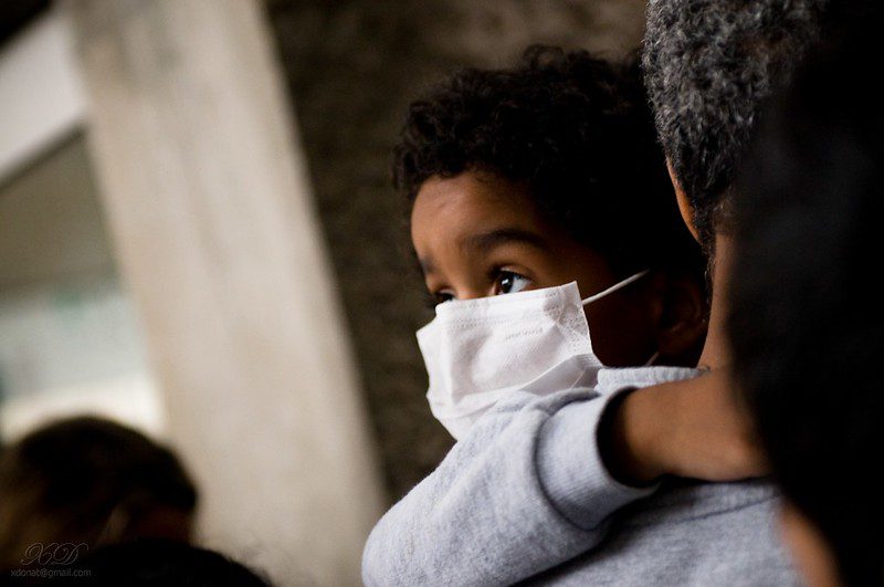 A child in a grey sweatshirt looks up over his white mask while being carried by an adult