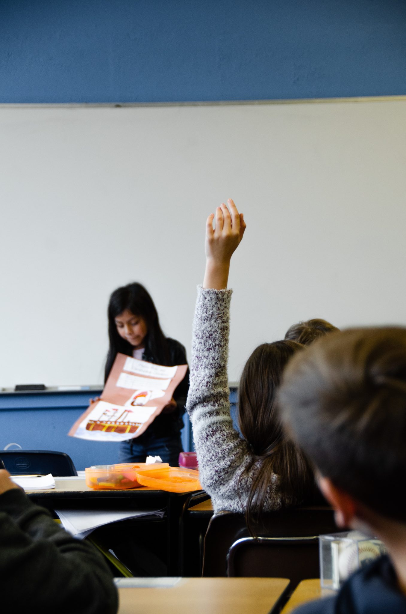 Student with her hand raised while a classmate shares a presentation