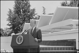 Black and white news photo of President Carter speaking in front of solar panels placed on the West Wing roof of the White House.