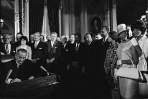 President Lyndon B. Johnson signs the Voting Rights Act of 1965 while Martin Luther King Jr. and others look on.