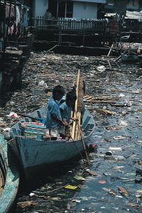 Two boys in a small rowboat look down at water that is heavily polluted by trash.
